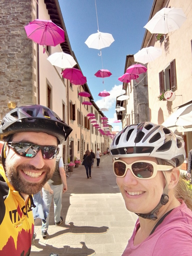 Two grinning bike tourists standing in front of Bagno di Romagna, still decorated with bright pink umbrellas in the sky for the Giro d'Italia.